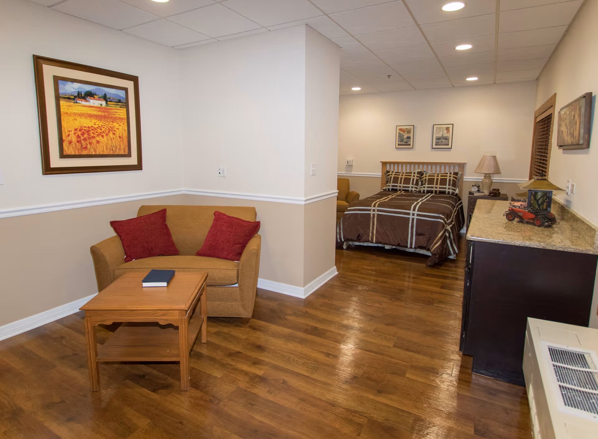 Interior view of a senior living facility room at Norwood Crossing featuring a wooden floor, a small sitting area with a brown loveseat and two red pillows, a wooden coffee table with a book on it, and a bedroom area with a bed covered in a brown plaid comforter. The room has beige walls with white trim, framed artwork on the walls, a dresser with decorative items, and recessed ceiling lights.