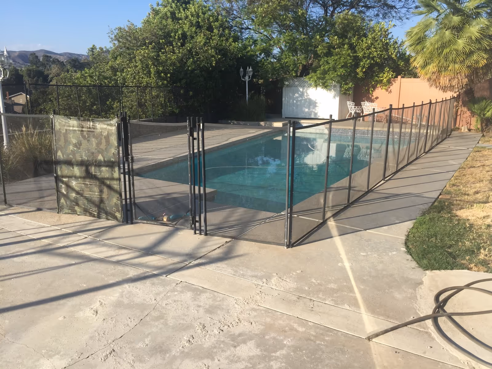 Outdoor swimming pool surrounded by a safety fence with a gate, set in a backyard with trees, a palm tree, and patio furniture in the background.