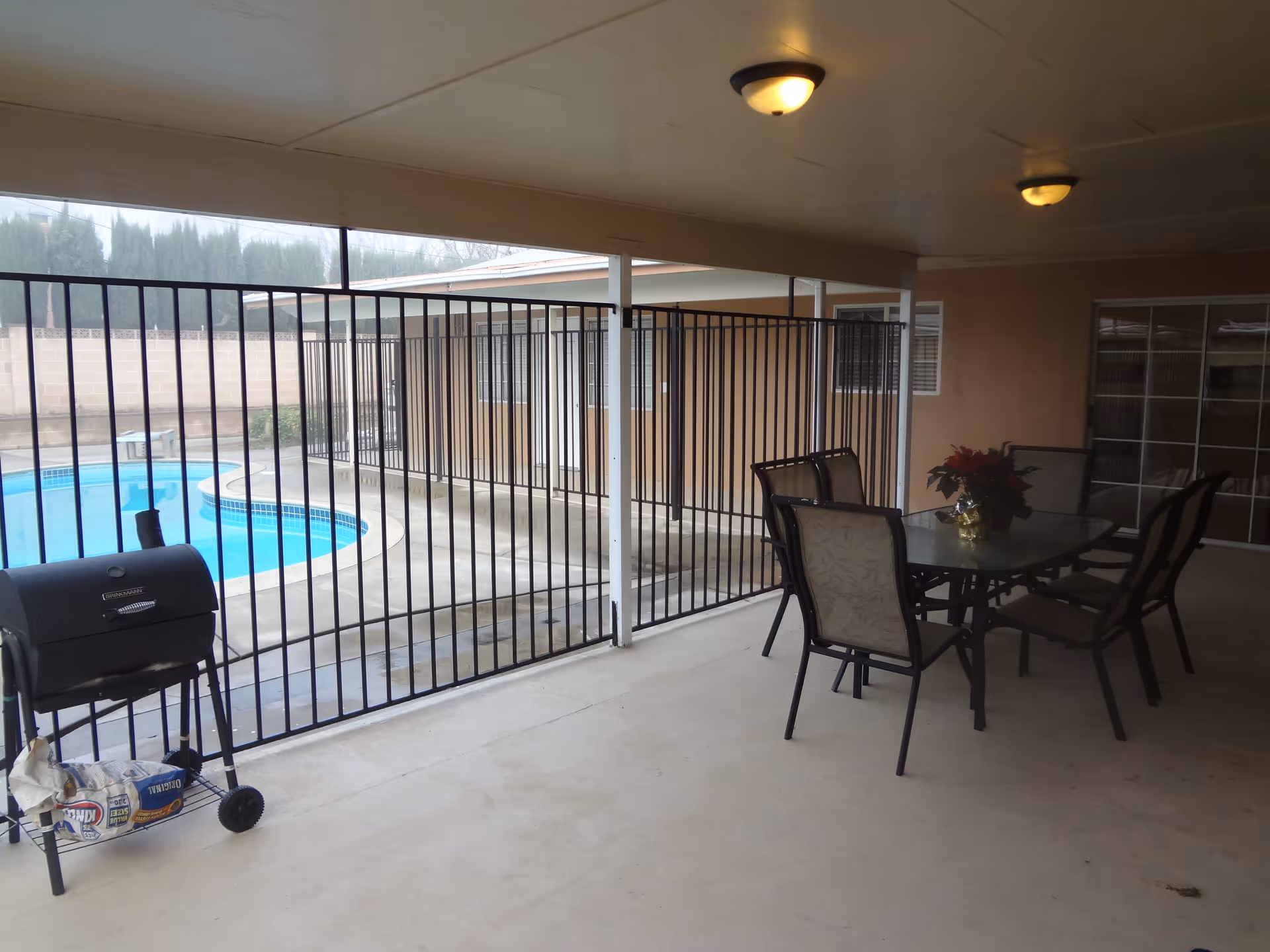 Covered patio area with a glass-top table and six chairs, a potted plant on the table, a charcoal grill with a bag of charcoal underneath, and a fenced swimming pool in the background.
