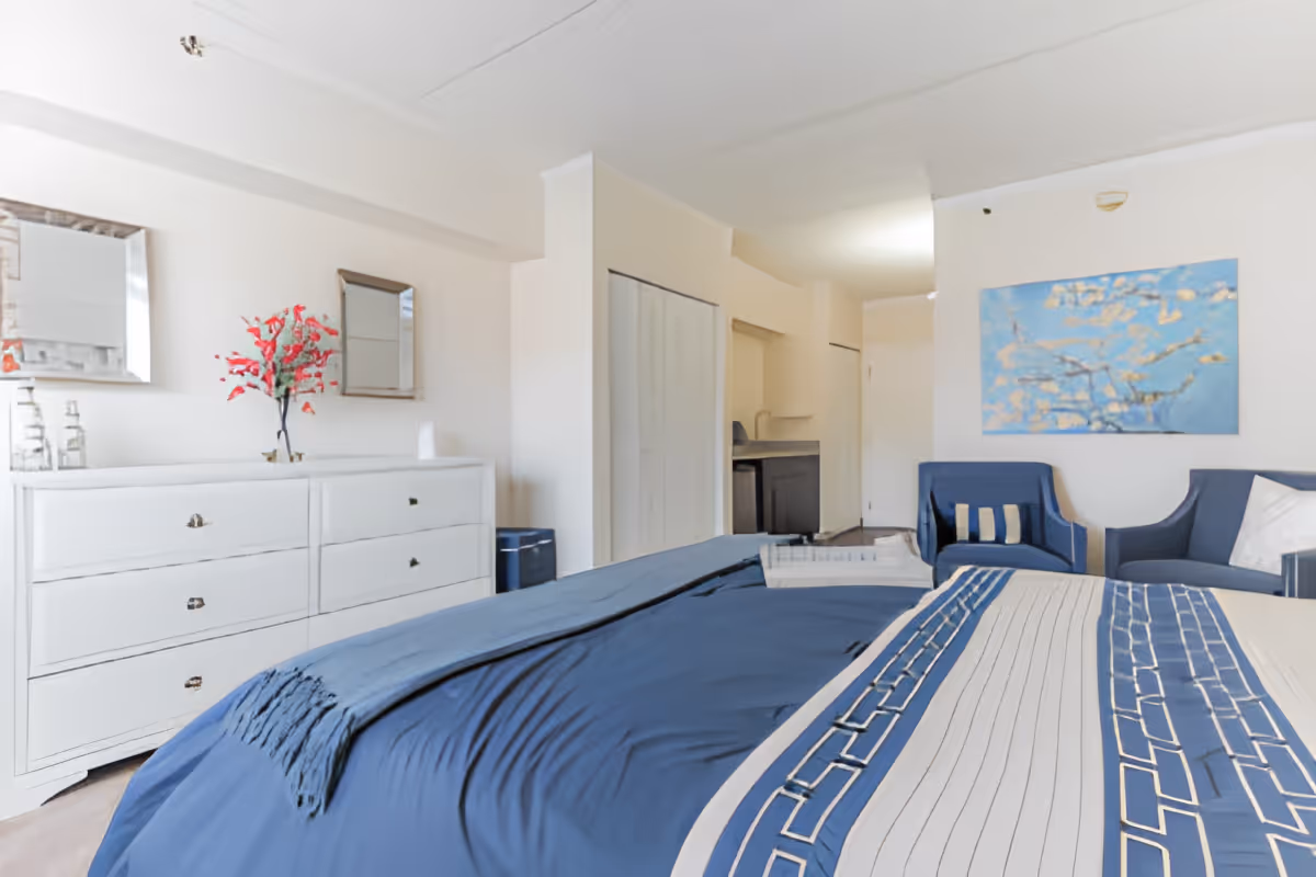 A bright bedroom featuring a bed with blue and white patterned bedding, a white dresser with a vase of red flowers and two mirrors above it, two blue armchairs with a decorative pillow, and a blue floral painting on the wall.