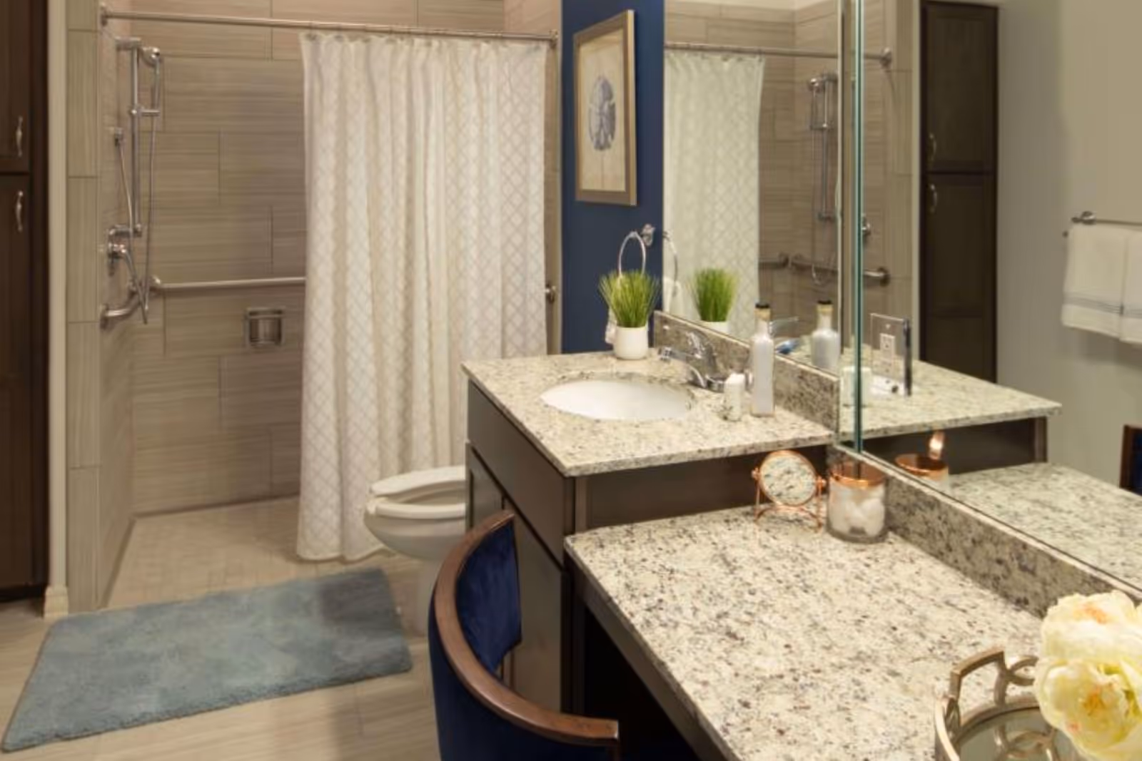 A modern bathroom with a granite countertop featuring a sink, decorative plants, and toiletries. There is a large mirror above the countertop. In the background, a shower area with a curtain and grab bars is visible, along with a toilet and a blue bath mat on the floor. A towel hangs on a rack on the wall.
