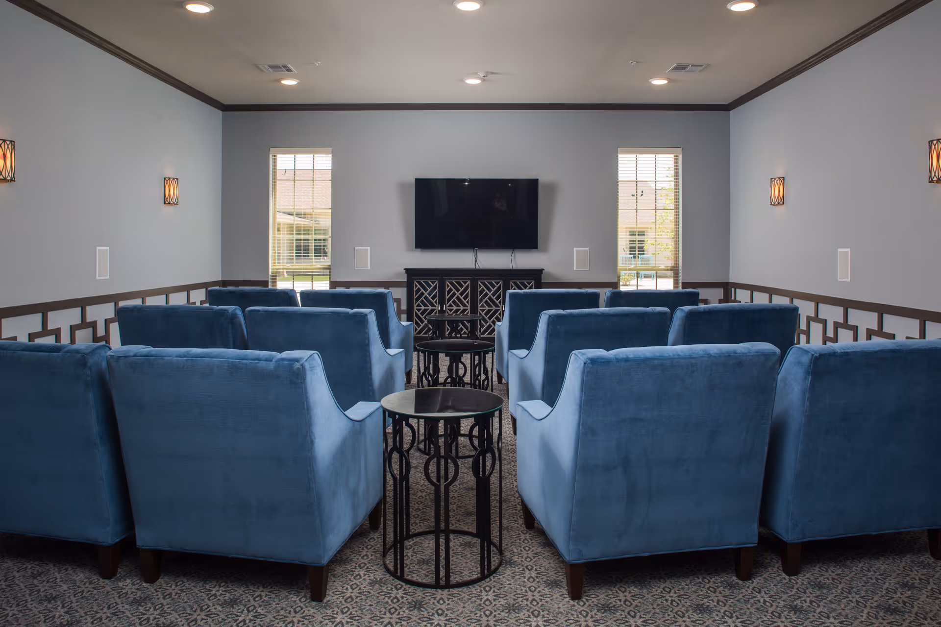 A small theater-style room with two rows of blue upholstered armchairs facing a wall-mounted flat screen TV. The room has two tall windows with blinds on either side of the TV, patterned carpet, and wall sconces providing soft lighting.