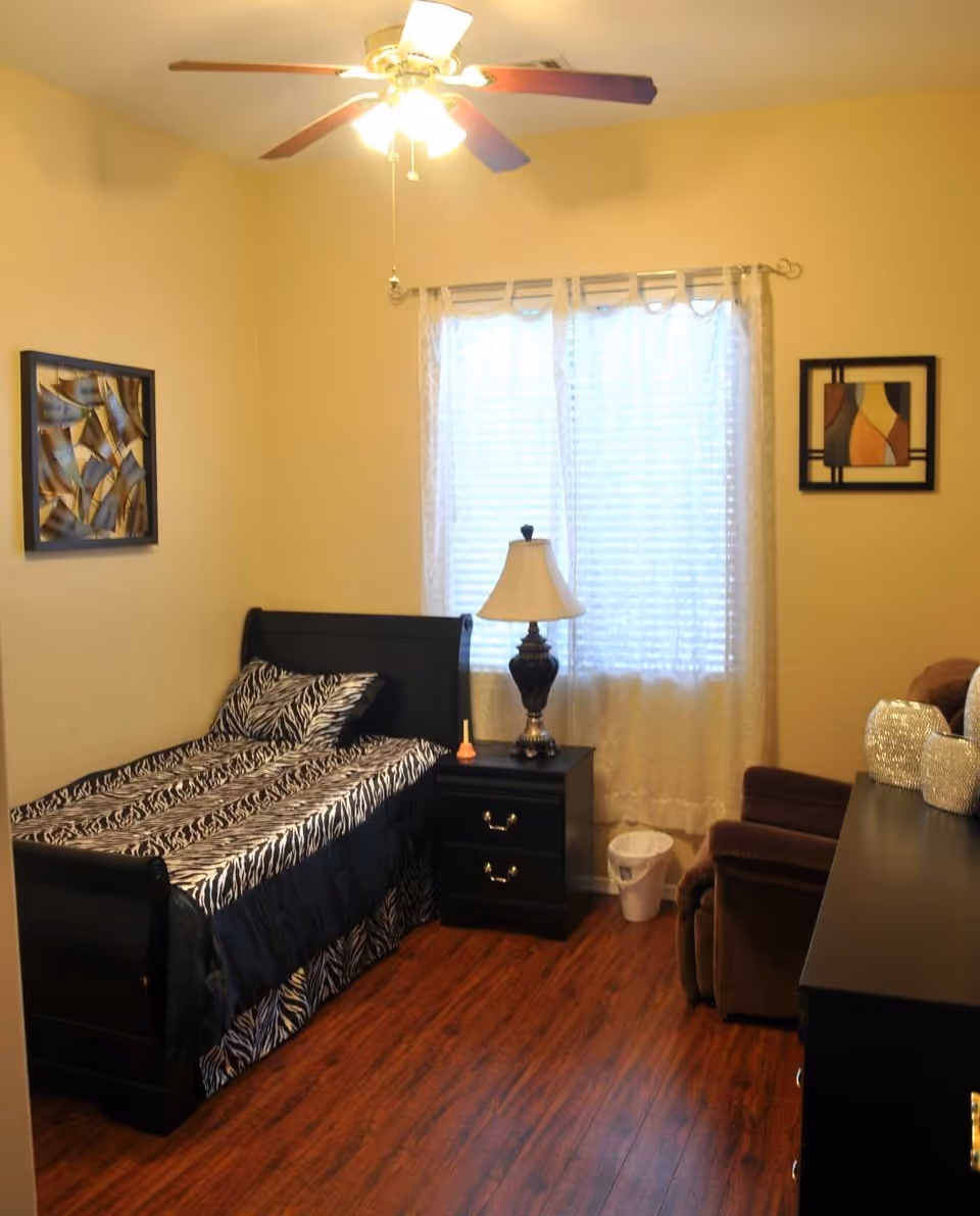 A small bedroom with a single bed featuring zebra print bedding, a black nightstand with a lamp, a brown recliner chair, a black dresser with decorative items, two framed abstract artworks on the walls, a window with white curtains, and a ceiling fan with lights.