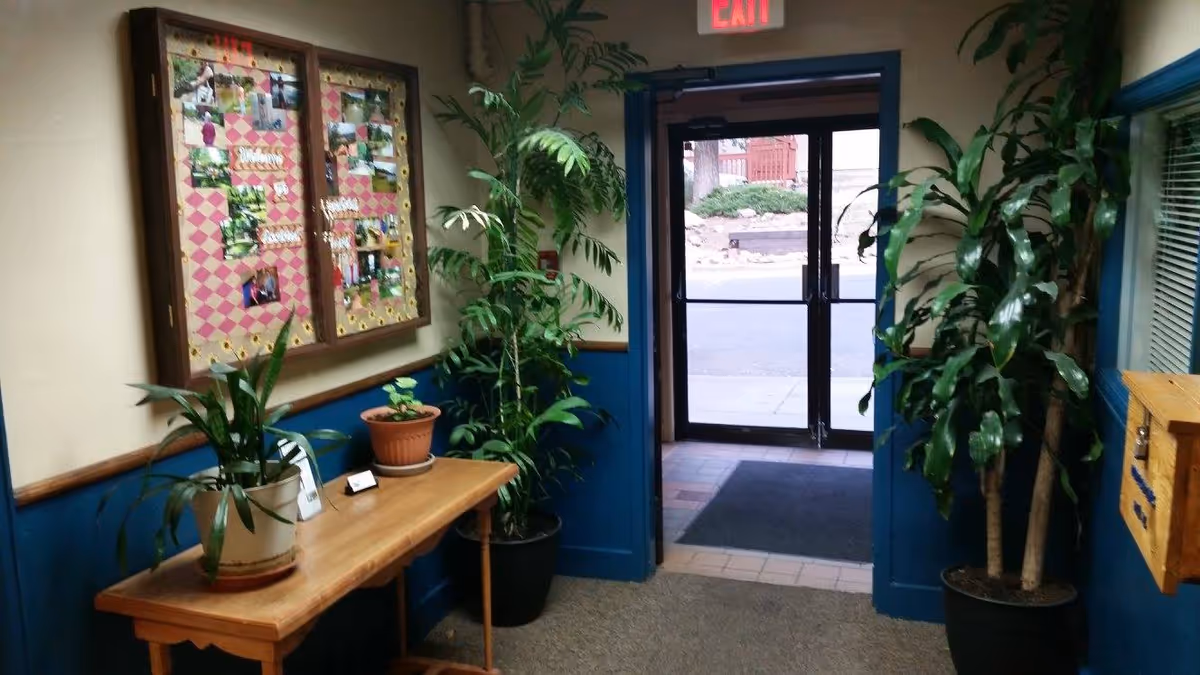 Interior view of a hallway in Brookside Assisted Living with a wooden table holding potted plants, a bulletin board with photos and notes on the wall, two large potted plants on either side of a glass exit door, and a wooden mailbox mounted on the wall.