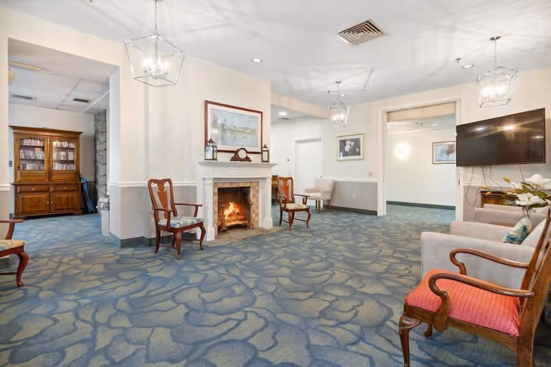 A cozy senior living common area with a lit fireplace, several wooden chairs with cushions, a wall-mounted flat screen TV, a wooden cabinet filled with DVDs, and soft blue patterned carpet. The room is softly lit with hanging lantern-style light fixtures and decorated with framed artwork on the walls.