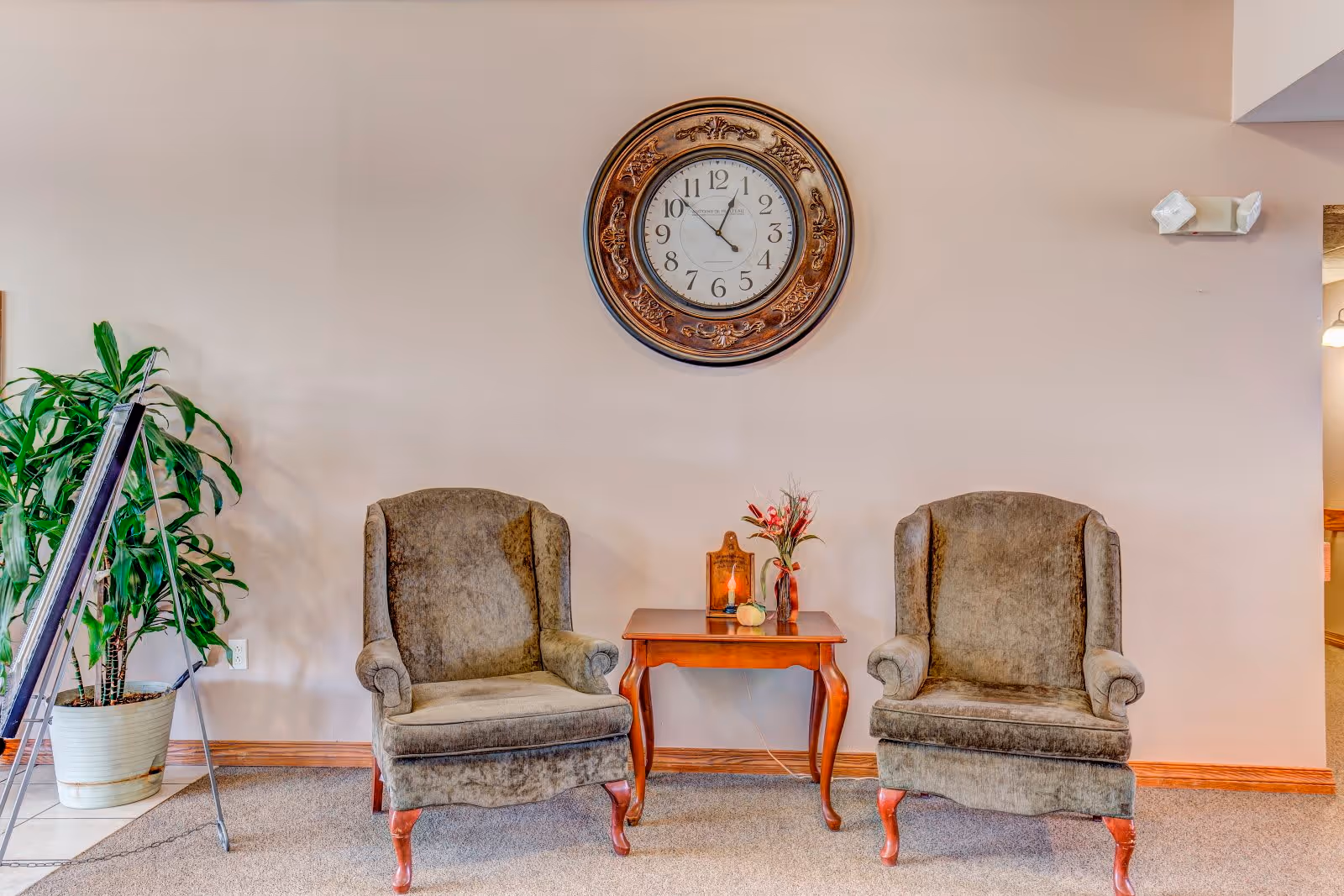 Two upholstered armchairs with wooden legs are positioned on either side of a small wooden table with a vase of flowers and a decorative item on it. Above the table, a large ornate wall clock is mounted on a beige wall. To the left, there is a tall green potted plant and part of a metal stand is visible. The floor is carpeted and the room has a calm, inviting atmosphere.