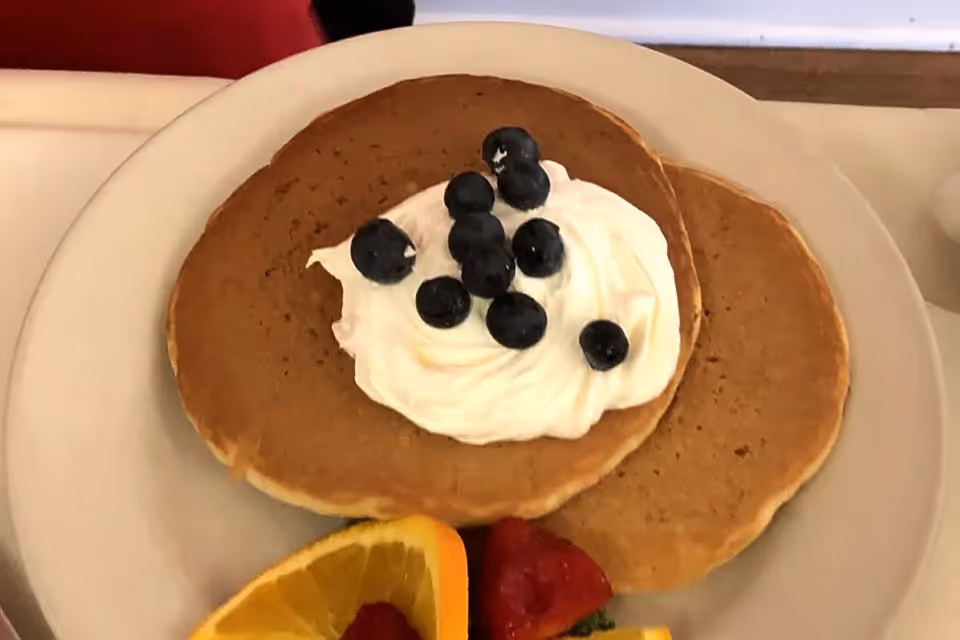 Two pancakes on a white plate topped with whipped cream and blueberries, accompanied by slices of orange and strawberries.