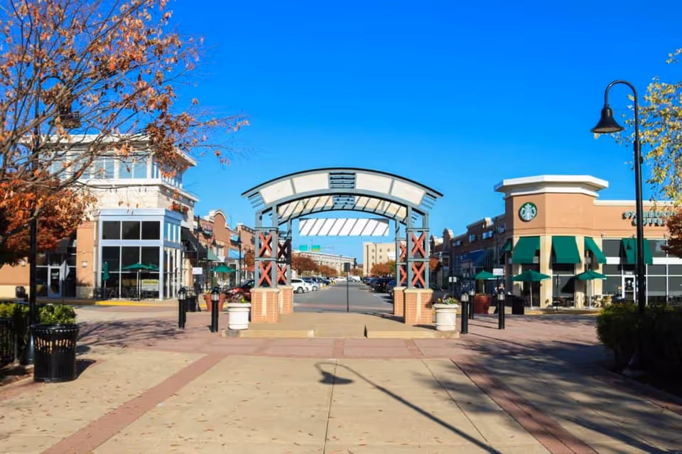 A bright, clear day view of a shopping plaza entrance with a decorative archway in the center. On the right side, there is a Starbucks with green awnings and outdoor seating. Trees with autumn-colored leaves line the walkway, and the sky is clear and blue.