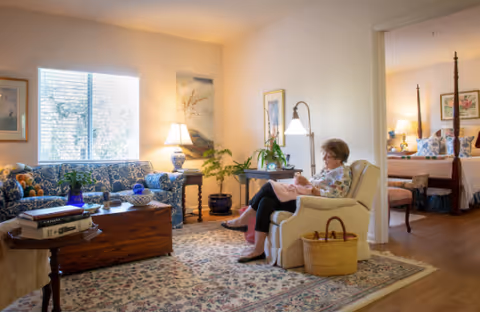 A cozy living room in Alderman Oaks Retirement Residence with a woman sitting in a beige armchair holding a baby. The room features a patterned rug, a wooden chest used as a coffee table, a blue patterned sofa, and several lamps providing warm lighting. There are framed pictures on the walls and a window letting in natural light. An adjoining bedroom with a four-poster bed is visible through an open doorway.