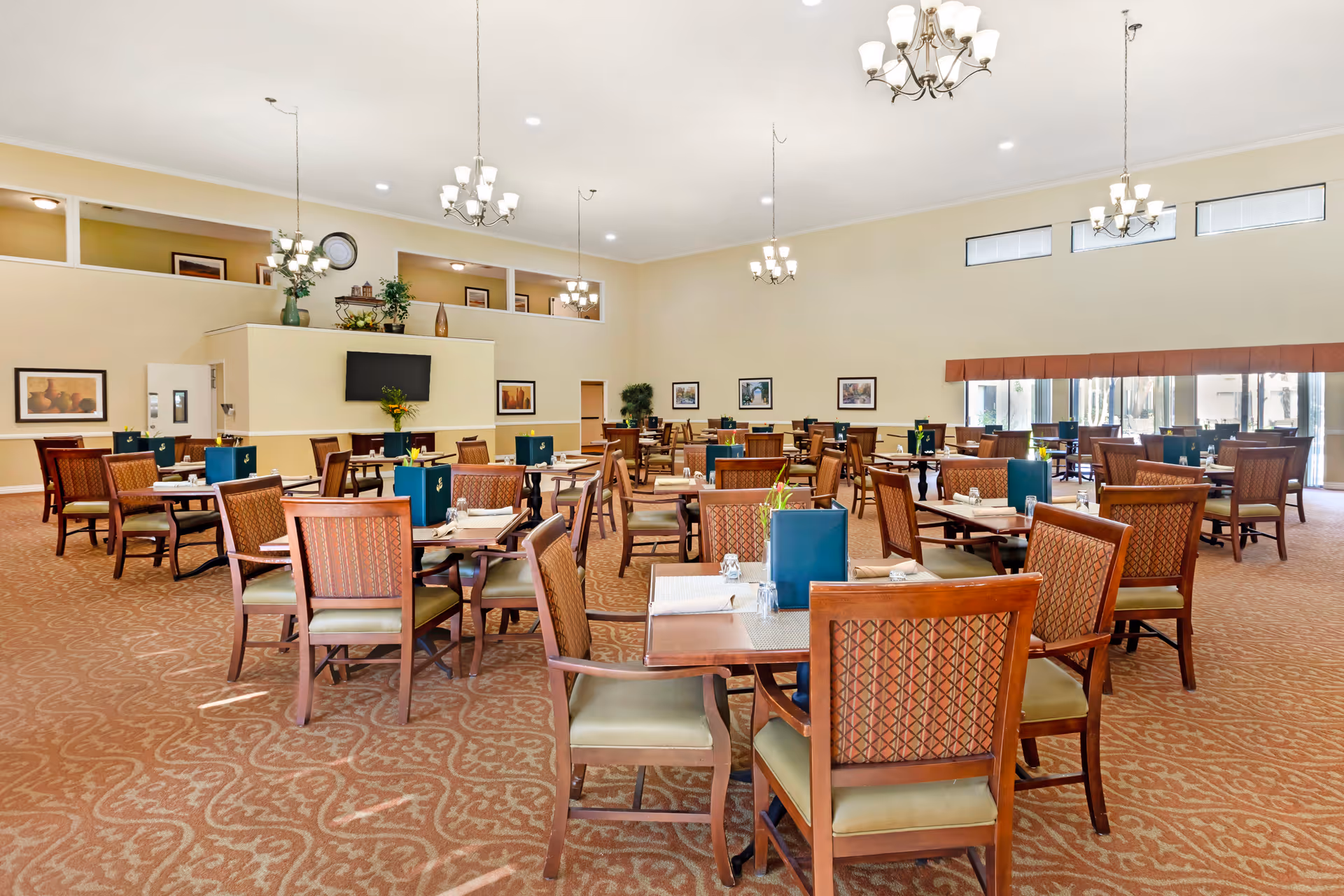 Spacious dining room with multiple wooden tables and chairs arranged neatly. Each table has a blue menu holder and a small flower vase. The room features beige walls, patterned carpet, several chandeliers hanging from the ceiling, and large windows with blinds allowing natural light to enter. There is a wall-mounted TV and framed artwork on the walls.
