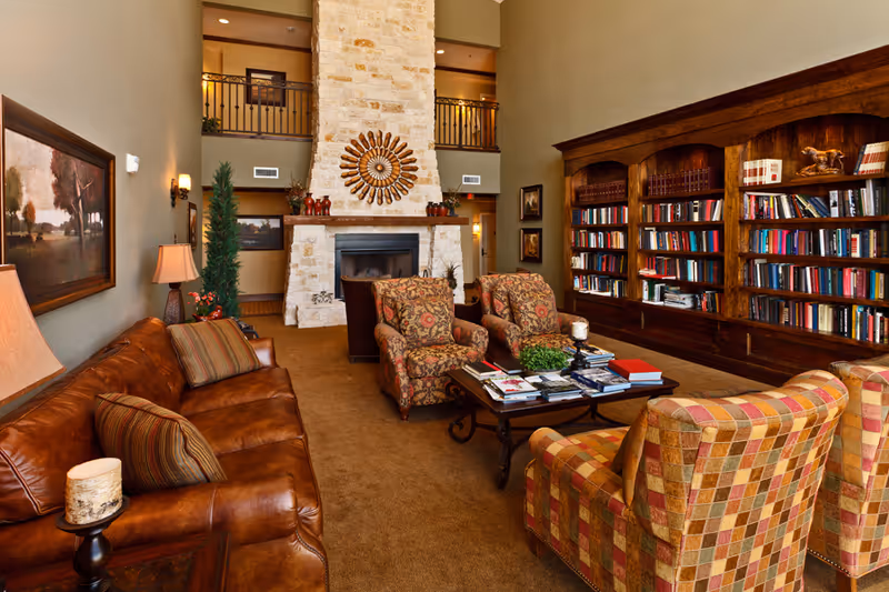 Cozy common room with leather sofa, patterned armchairs, a coffee table, large stone fireplace and a wall of bookshelves.