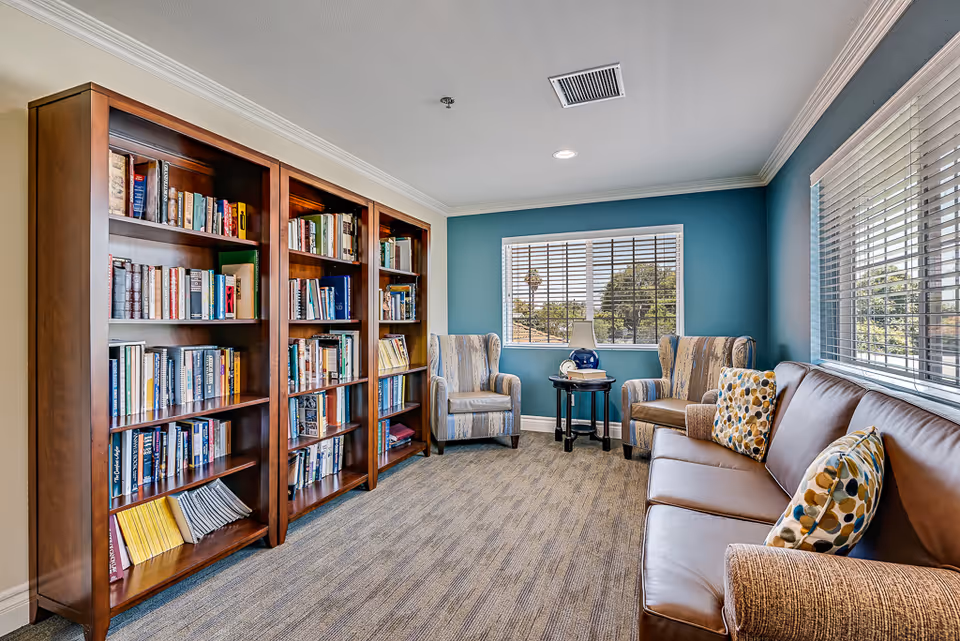 A bright seating and reading room with tall wooden bookshelves, upholstered armchairs around a small table, and a leather sofa by windows.