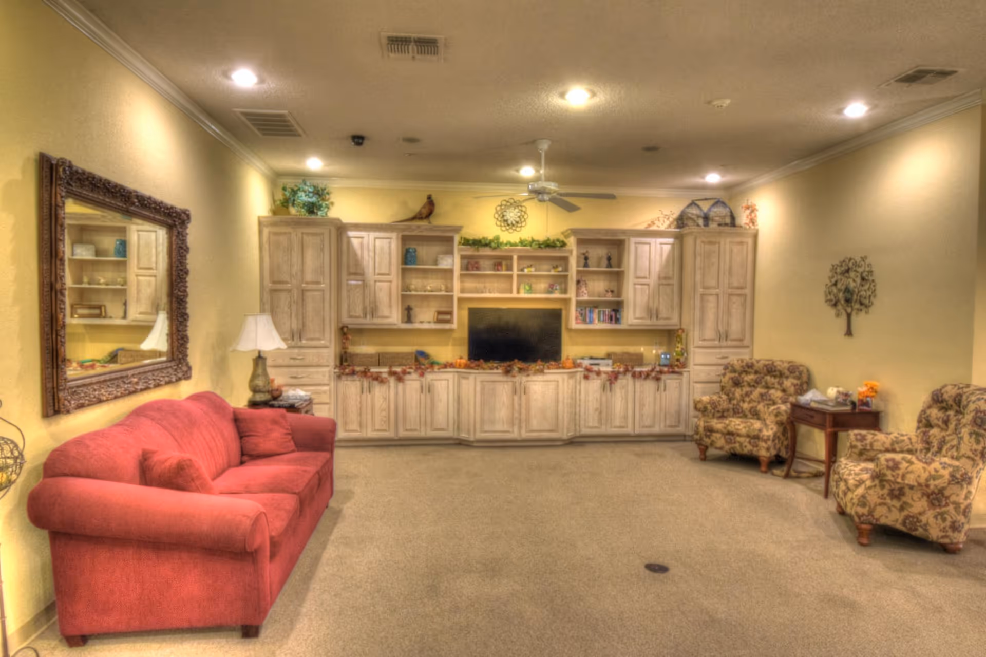 A cozy living room with a red sofa on the left, two floral-patterned armchairs on the right, and a wooden entertainment center with shelves and cabinets along the back wall. The entertainment center holds a flat-screen TV and decorative items. The walls are painted light yellow, and there is a large ornate mirror above the sofa. The room is softly lit with ceiling lights and a table lamp beside the sofa.
