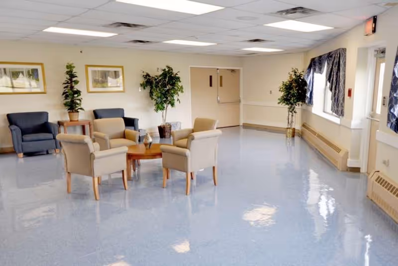A clean and bright common area with a shiny blue floor, featuring a seating arrangement of four beige armchairs around a small wooden table, two dark blue armchairs against the wall, potted plants, framed pictures on the wall, and windows with blue curtains.