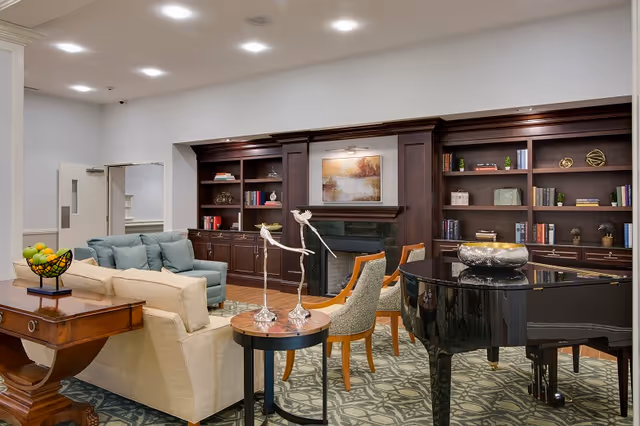 Well-lit communal living room with sofas, chairs, a grand piano, built-in wood bookshelves and a fireplace.