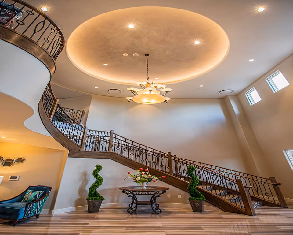 Spacious foyer with a sweeping curved staircase, a central chandelier above a round table with flowers and topiary plants.