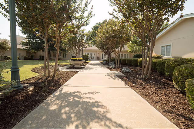 A paved walkway lined with trees and shrubs leading to the entrance of a single-story building under a clear sky, with well-maintained landscaping on both sides.