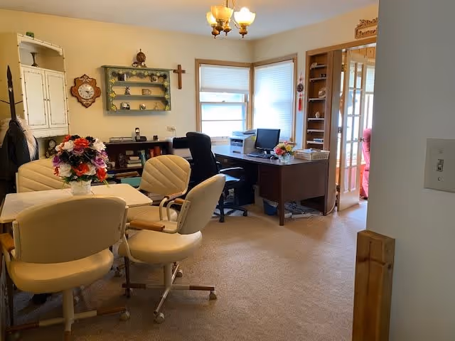 A cozy interior room with beige carpet and walls, featuring a round table with four cream-colored chairs and a vase of colorful flowers on top. In the background, there is a wooden desk with a computer, office chair, and printer near two windows with blinds. The room also has decorative shelves, a wall clock, a cross, and a chandelier light fixture. A doorway with glass panels leads to another room with a pink chair visible.