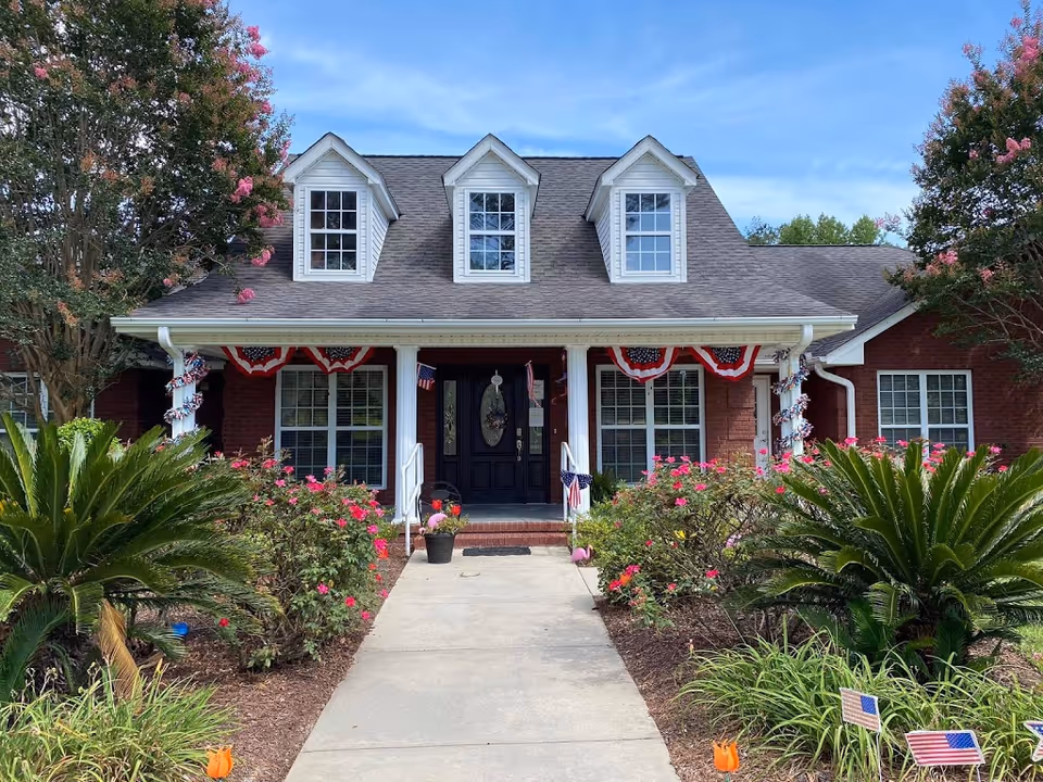 Front exterior view of a single-story brick building with a dark shingled roof and three dormer windows. The entrance has a covered porch supported by white columns, decorated with red, white, and blue patriotic bunting and small American flags. There are well-maintained flower beds with pink flowers and green shrubs lining the concrete walkway leading to the black front door.