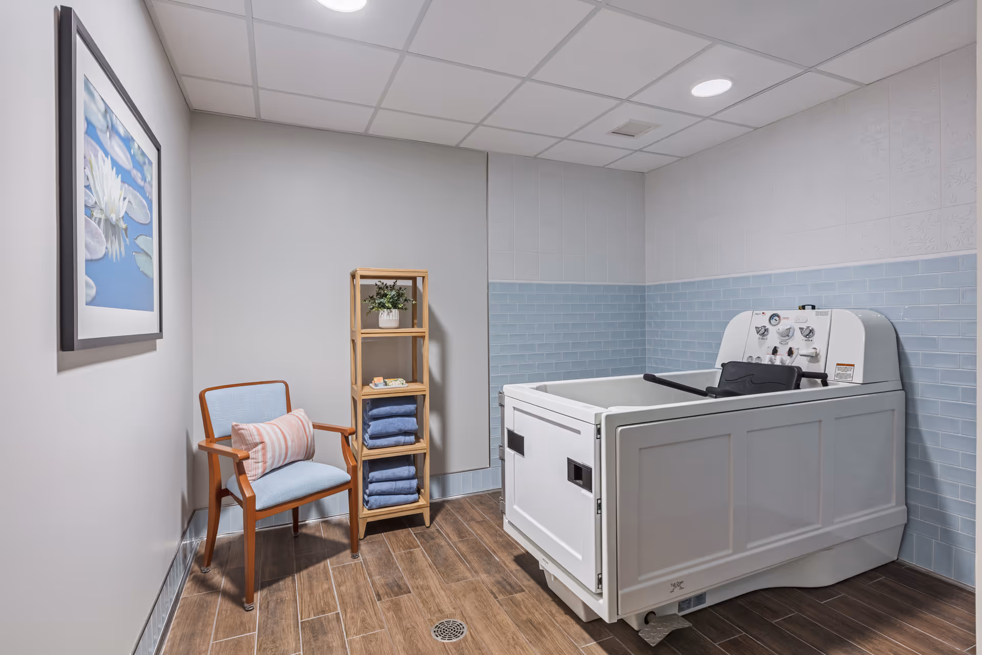A clean and well-lit bathing room with a large white assisted bathtub on the right, a wooden chair with a blue cushion and a striped pillow on the left, and a wooden shelving unit holding folded blue towels and a small plant. The walls are partly tiled with light blue subway tiles and the floor has wood-look tiles. A framed picture of water lilies hangs on the left wall.