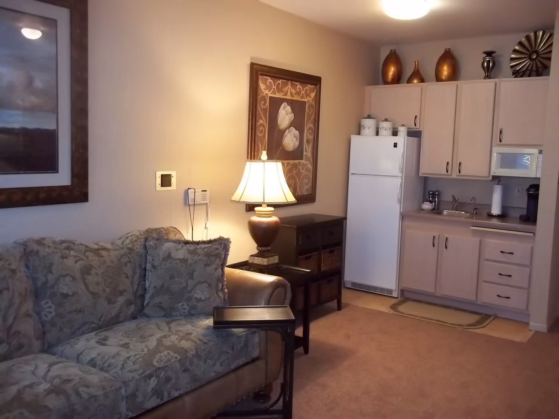 A cozy living area with a patterned sofa and a side table with a lamp. Adjacent to the living space is a small kitchen area with light wood cabinets, a white refrigerator, a microwave, and a sink. Decorative vases and canisters are placed on top of the cabinets. The walls are adorned with framed artwork.