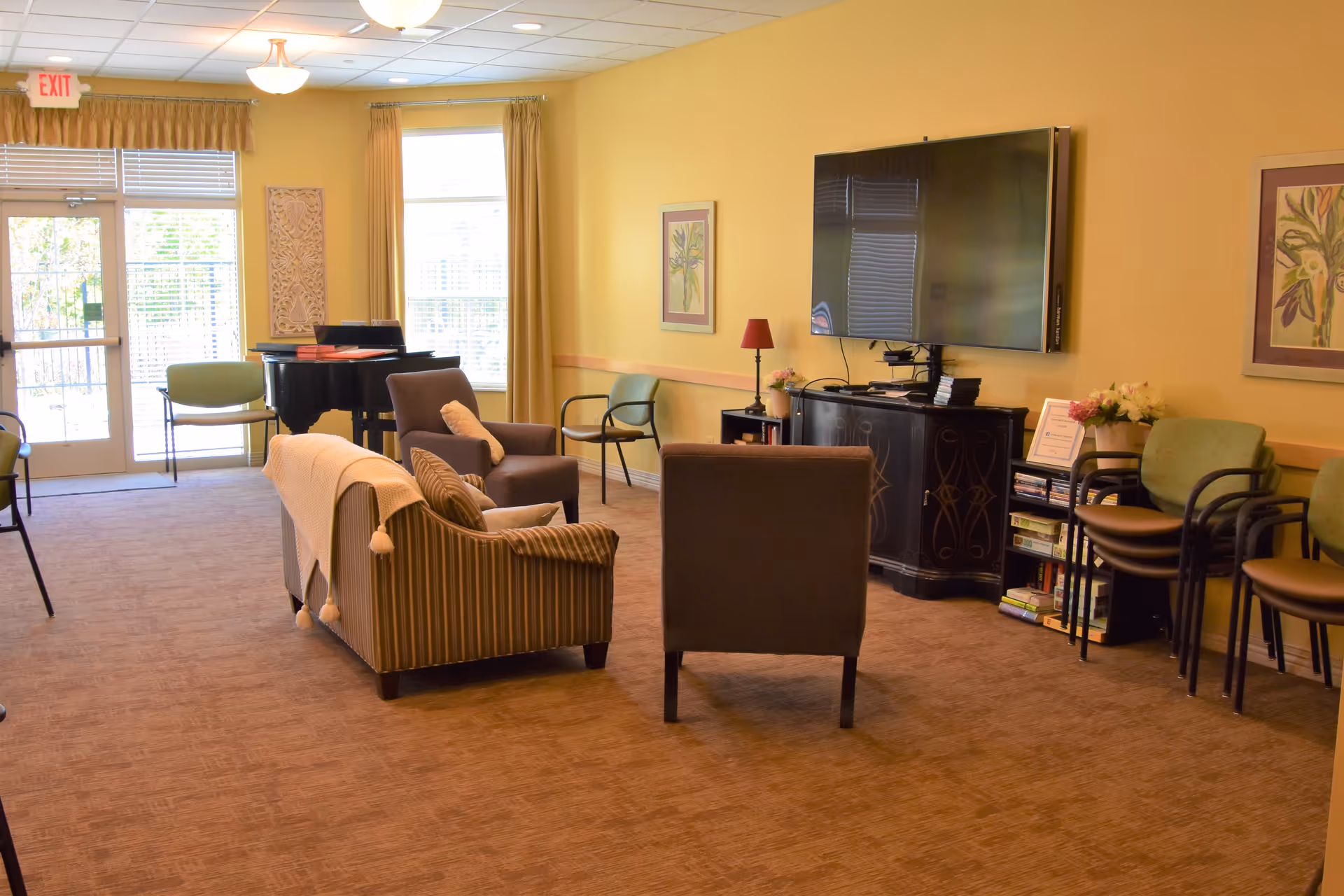 A cozy living room area in Stone Creek Assisted Living and Memory Care with a striped sofa, two armchairs, a large flat-screen TV mounted on a yellow wall, and a black cabinet below the TV. There are framed floral artworks on the walls, a small table with a lamp and flowers, several stacked chairs, a piano near a window, and a glass door leading outside.