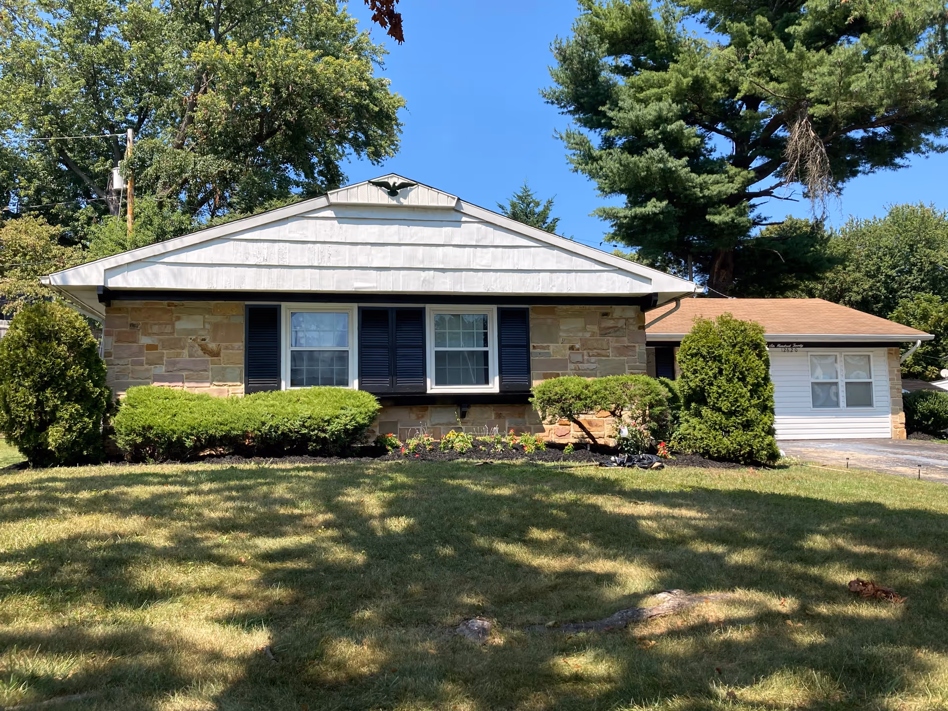 Front exterior of a single-story stone and siding house with lawn, shrubs, and trees under a clear blue sky.