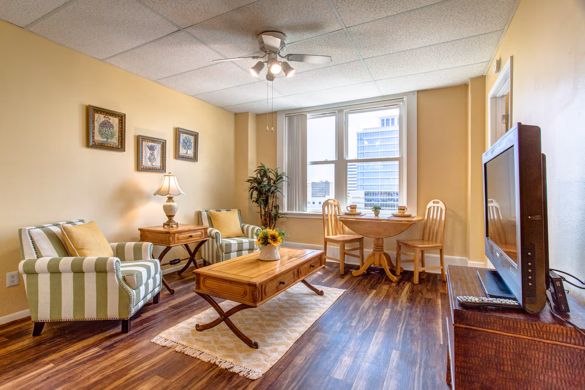 A cozy living room with two striped armchairs with yellow cushions, a wooden coffee table with a vase of sunflowers, a side table with a lamp, a small round dining table with two wooden chairs near a window showing a cityscape, and a flat-screen TV on a wooden stand.