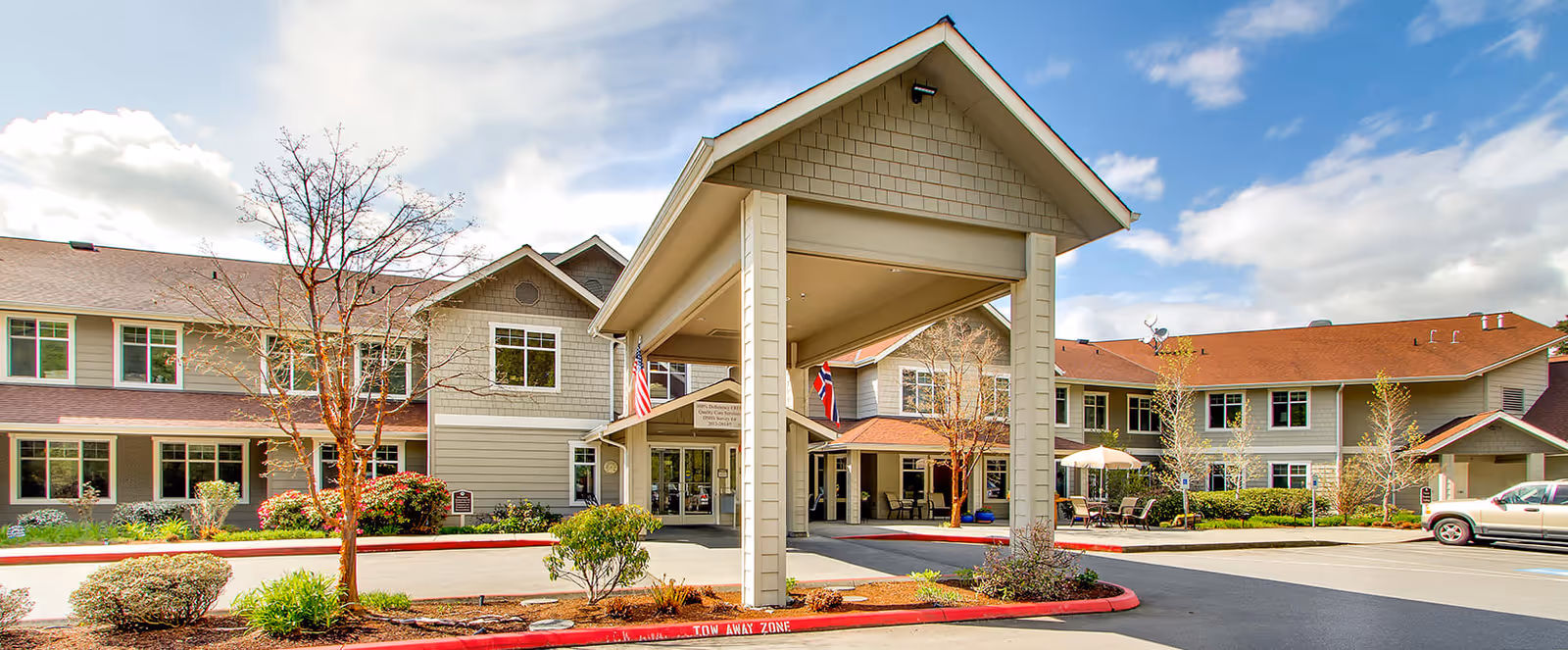 Front exterior view of Brookdale Montclair Poulsbo senior living facility with a covered entrance, landscaped bushes and trees, and a clear sky with some clouds.