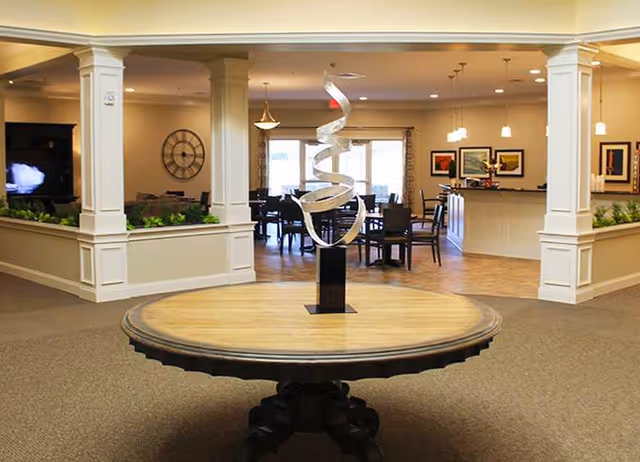 Interior view of a senior living facility featuring a round wooden table with a modern spiral metal sculpture in the center. The background shows a dining area with tables and chairs, pendant lights, framed artwork on the walls, and large windows letting in natural light.