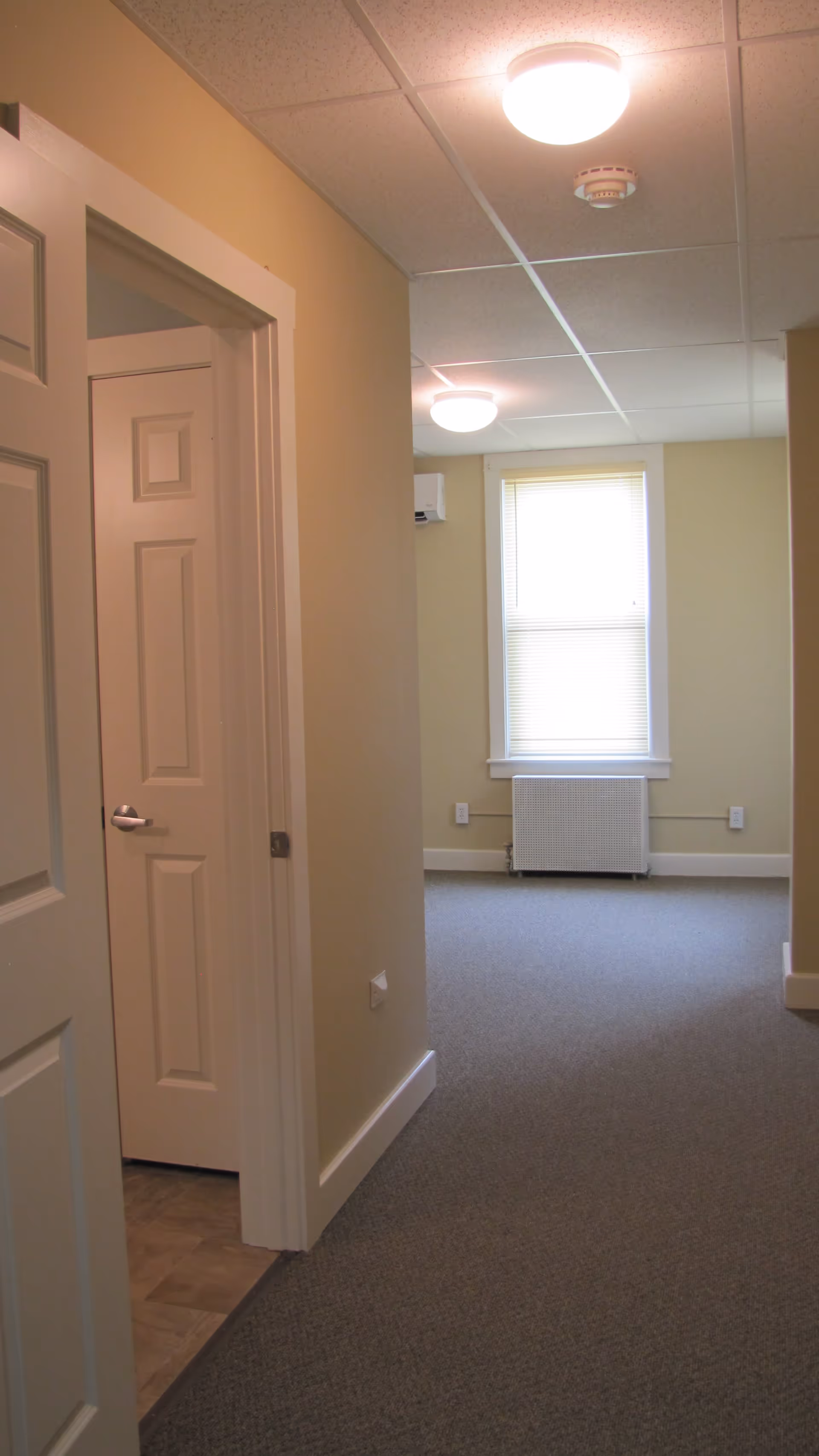 Interior view of a hallway in a senior living facility with beige walls, carpeted floor, and a window at the end with blinds partially closed. There are two closed white doors on the left side and ceiling lights illuminating the space.