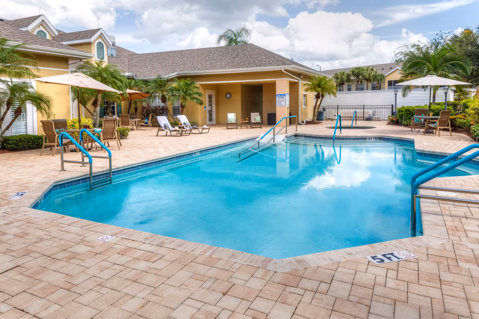 Outdoor swimming pool area at a senior living facility with lounge chairs, tables with umbrellas, palm trees, and a building in the background under a partly cloudy sky.