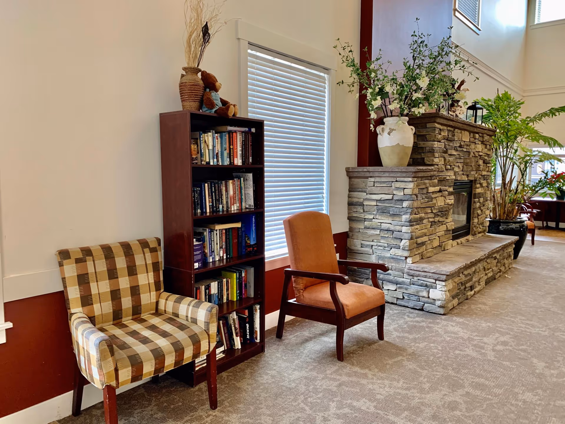 A cozy sitting area in a senior living facility featuring a plaid armchair, an orange cushioned wooden chair, a tall bookshelf filled with books, and a stone fireplace decorated with a large vase of flowers. The room has carpeted flooring and large windows with blinds allowing natural light to enter.