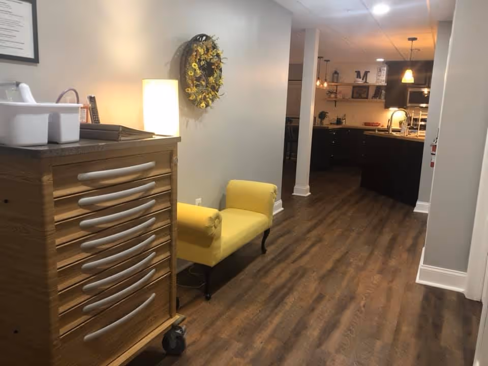 Interior view of a senior living facility hallway with wooden flooring, a yellow upholstered bench against a light gray wall, a wooden cabinet with multiple drawers on wheels, a lamp, and a decorative wreath on the wall. The hallway leads to a kitchen area with dark cabinetry, a sink, and pendant lights.