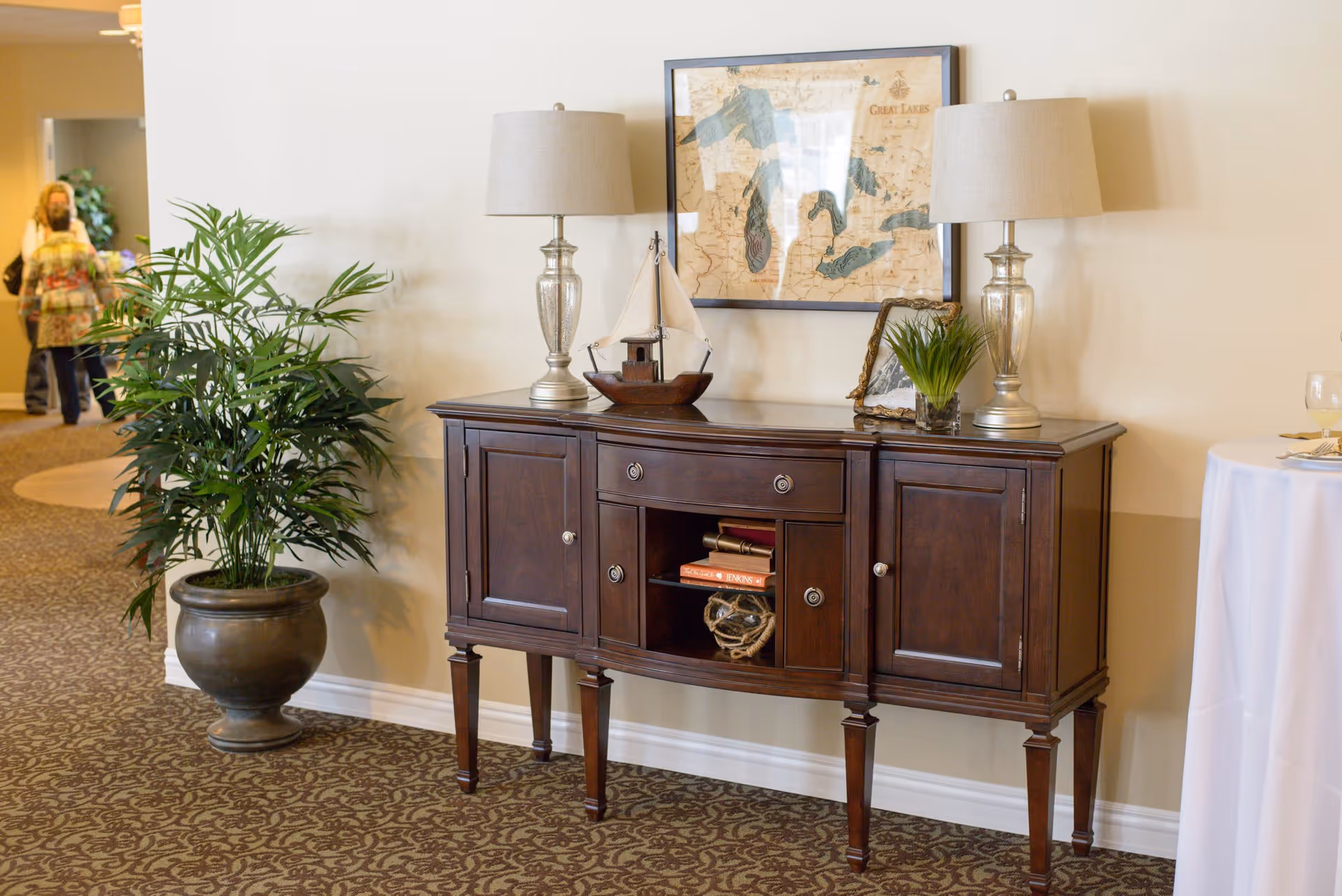 A wooden sideboard with two table lamps, a decorative sailboat, a small plant, and books on it, placed against a beige wall with a framed map of the Great Lakes above it. To the left, there is a large potted plant, and in the background, two people are walking in a hallway. On the right side, a round table with a white tablecloth and a glass of wine is partially visible.