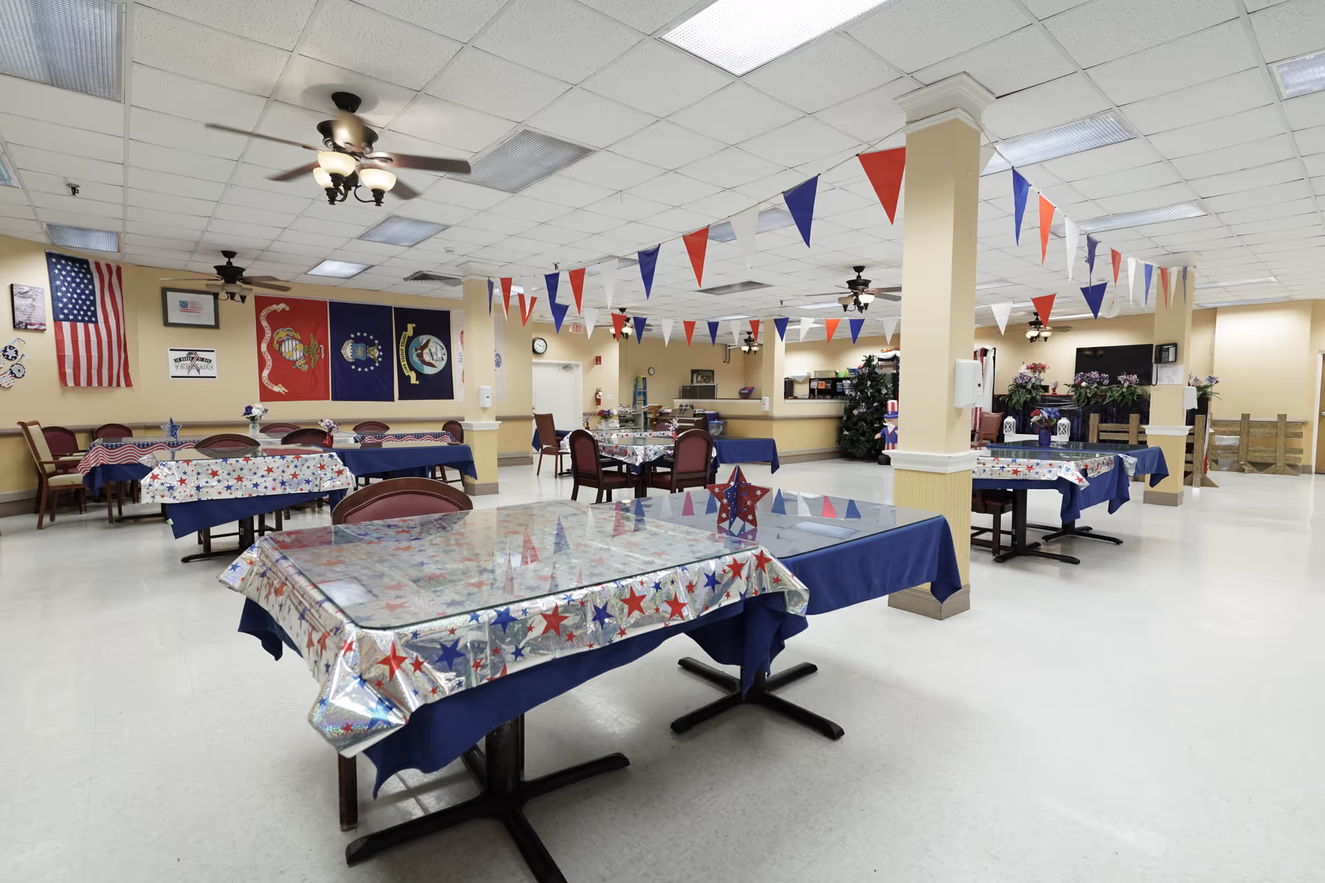 A large dining room decorated with red, white, and blue patriotic bunting and tablecloths. Several tables and chairs are arranged throughout the room. Flags and banners hang on the far wall, including the American flag and military branch flags. Ceiling fans and fluorescent lights are visible on the ceiling.
