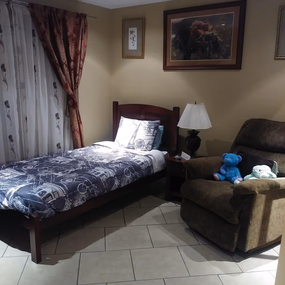 A cozy bedroom with a single bed, nightstand and lamp next to a recliner holding stuffed animals.