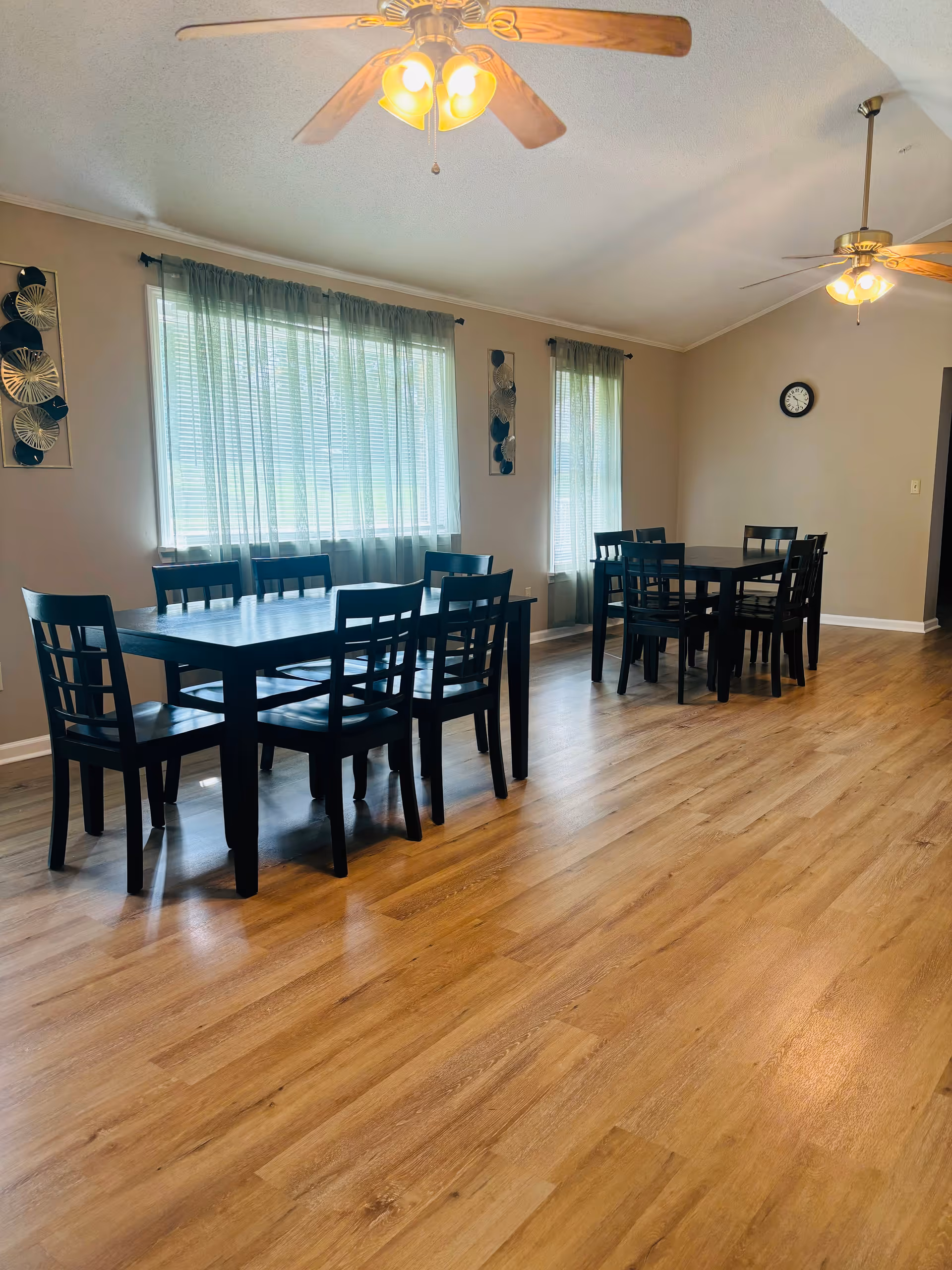 A dining room with two black rectangular tables, each surrounded by six matching black chairs. The room has light brown wooden flooring, beige walls, two ceiling fans with lights, and two windows with sheer green curtains allowing natural light to enter. A clock is mounted on the wall.