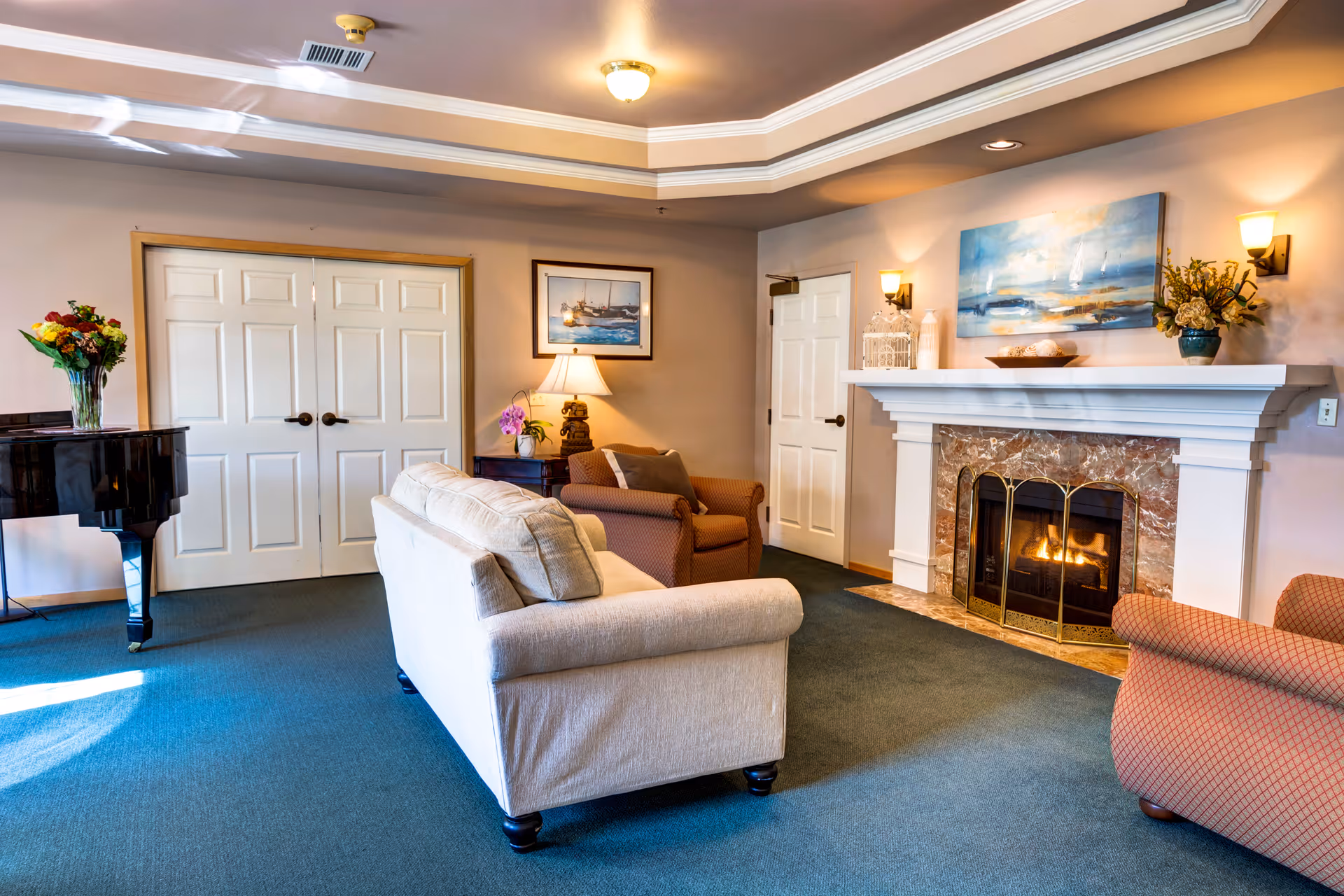 A cozy living room with a beige sofa and two armchairs arranged around a fireplace with a marble surround and a fire burning inside. Above the fireplace is a large painting of a seascape. The room has a blue carpet, a grand piano with a vase of flowers on top, and a side table with a lamp and decorative items. There are double white doors and a single white door in the background.