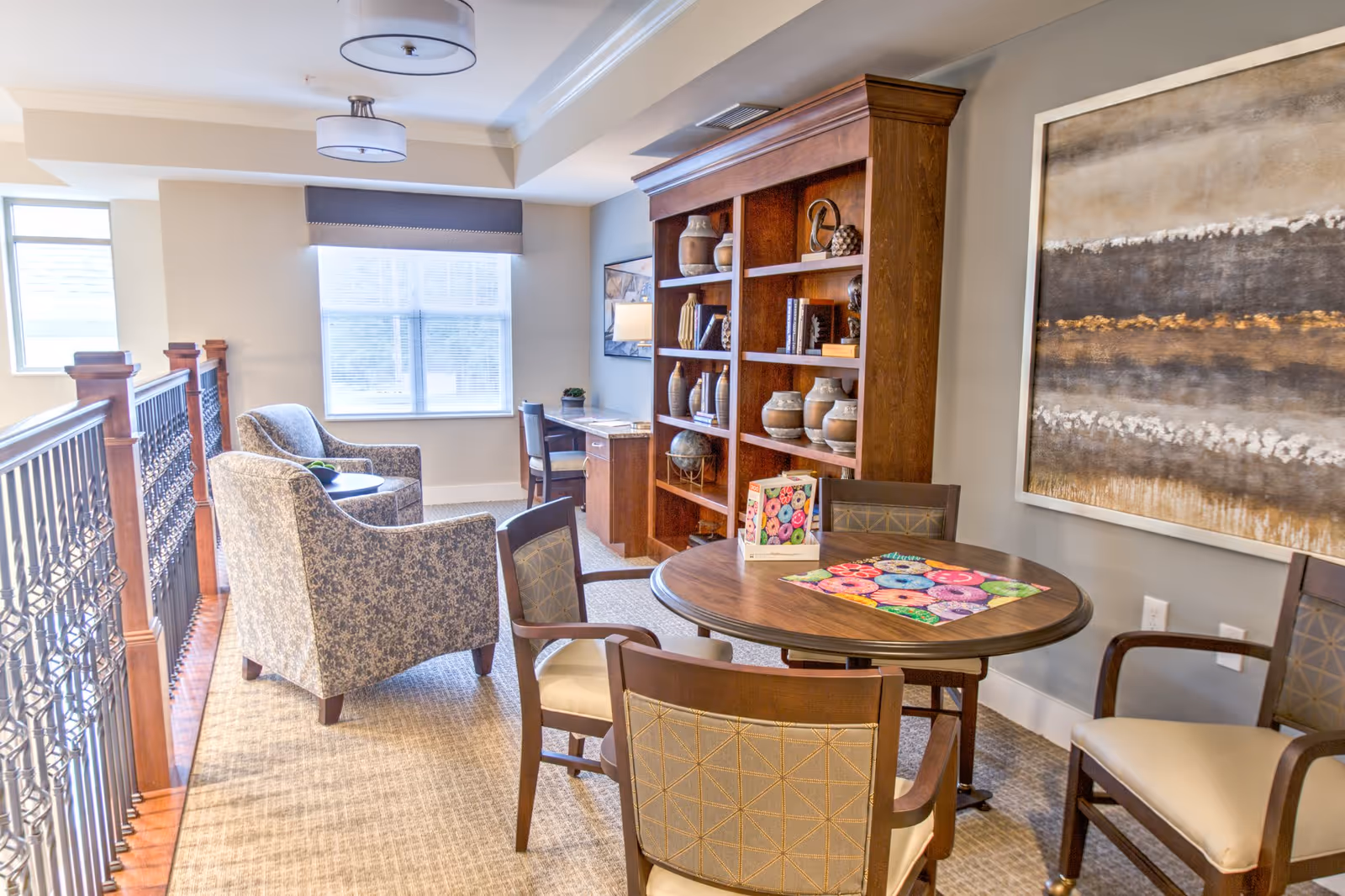 Cozy senior living common area with a round table and chairs, upholstered armchairs by a window, and a wooden bookshelf with decorative items.