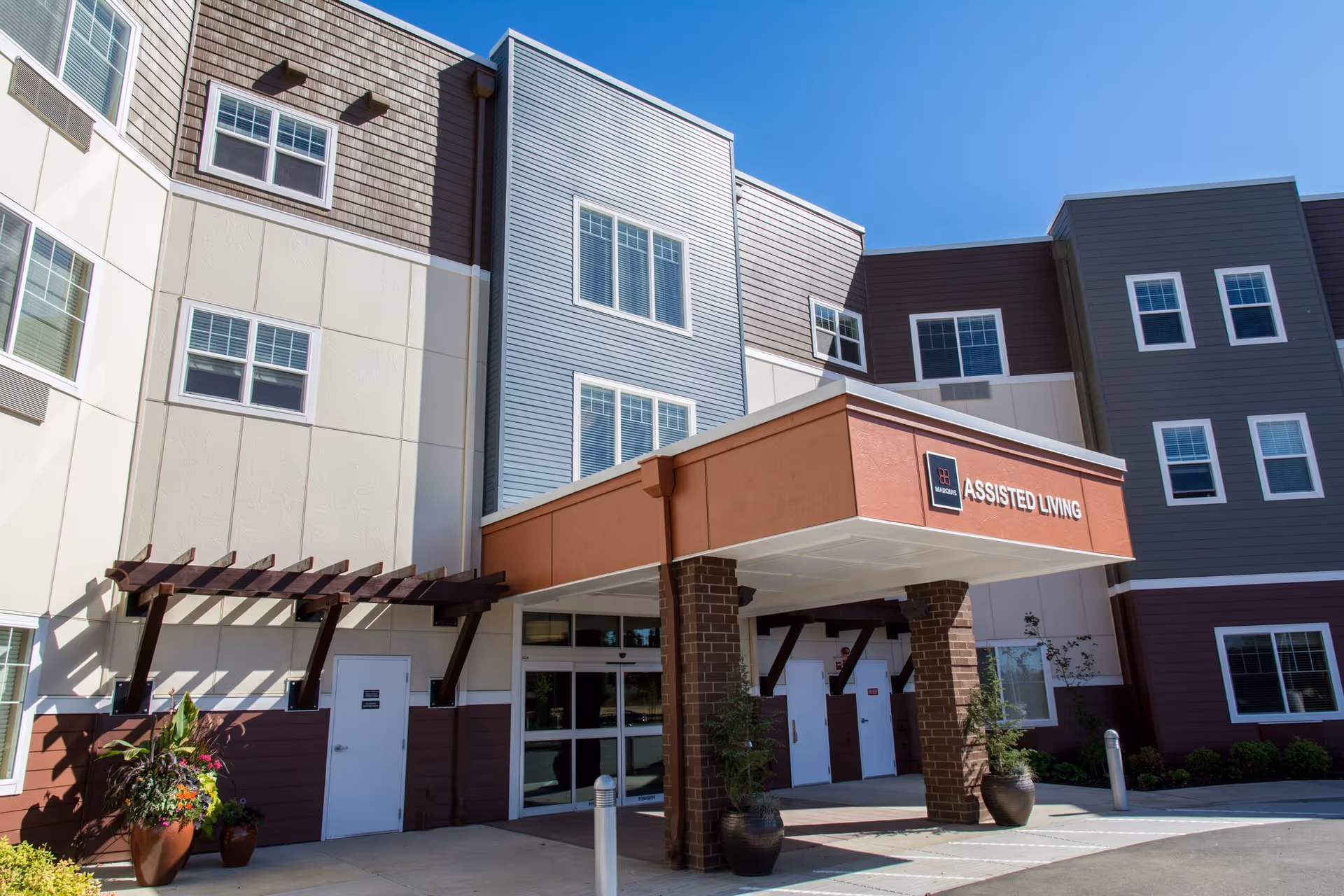 Front entrance of an assisted living building with a covered portico, potted plants and multi-story siding under a clear blue sky.