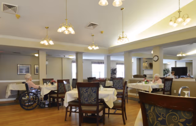 A dining room in a senior living facility with several tables covered with white tablecloths and chairs around them. Two elderly women are seated, one in a wheelchair and another at a table. The room has wooden flooring, multiple ceiling light fixtures, and large windows letting in natural light.