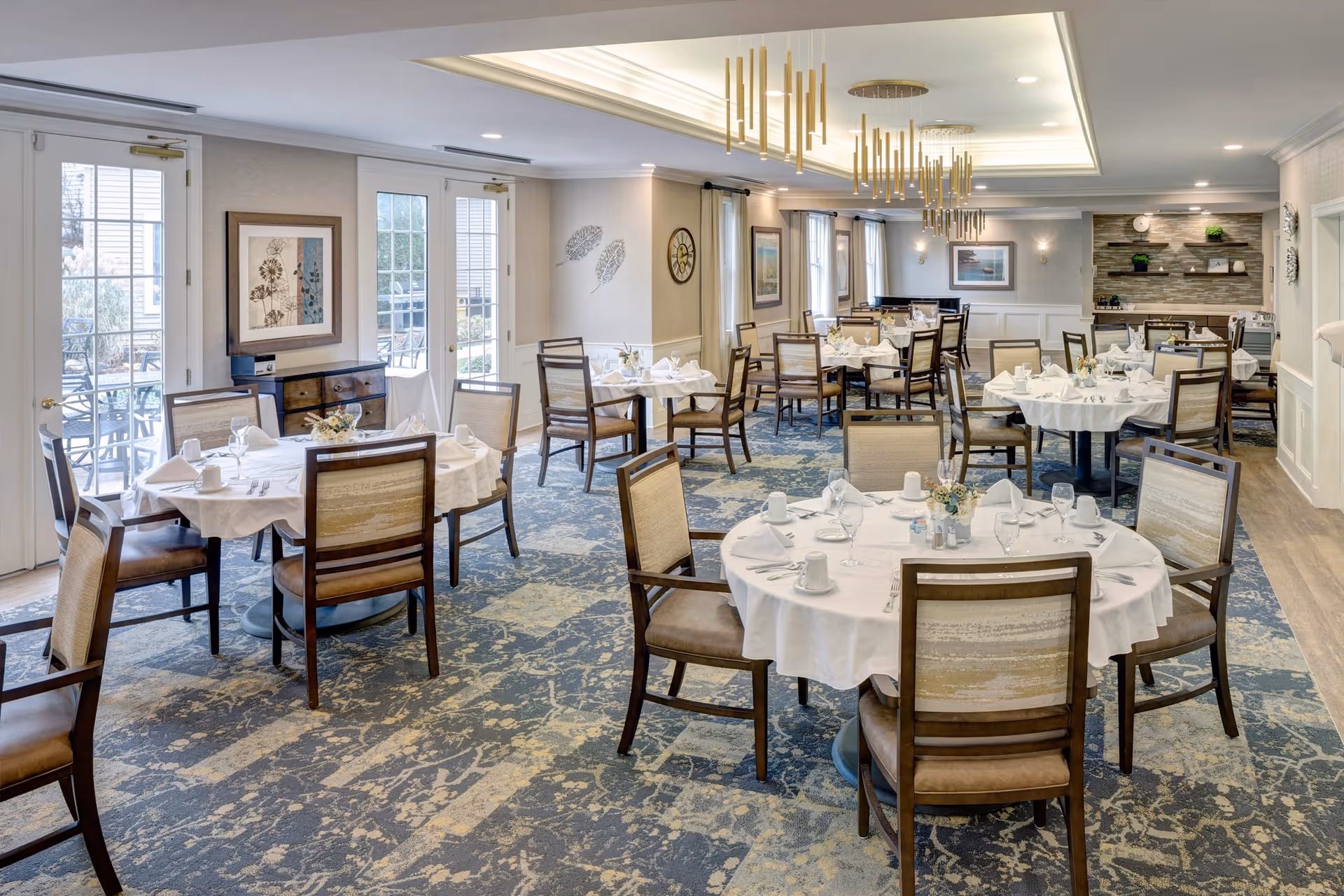 A spacious dining room with multiple round tables covered with white tablecloths, each set with napkins, cups, glasses, and small floral centerpieces. The room has large windows and glass doors letting in natural light, modern chandeliers hanging from the ceiling, and a patterned carpet. The walls are decorated with framed artwork and wall-mounted shelves with plants and decor items.
