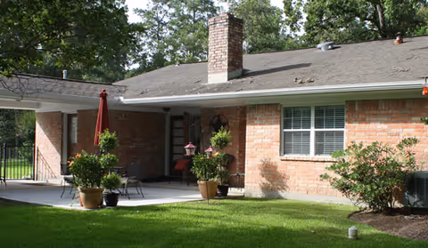 Exterior view of a single-story brick building with a covered patio area. The patio has outdoor furniture including a table, chairs, and potted plants. There is a green lawn in the foreground and trees in the background.