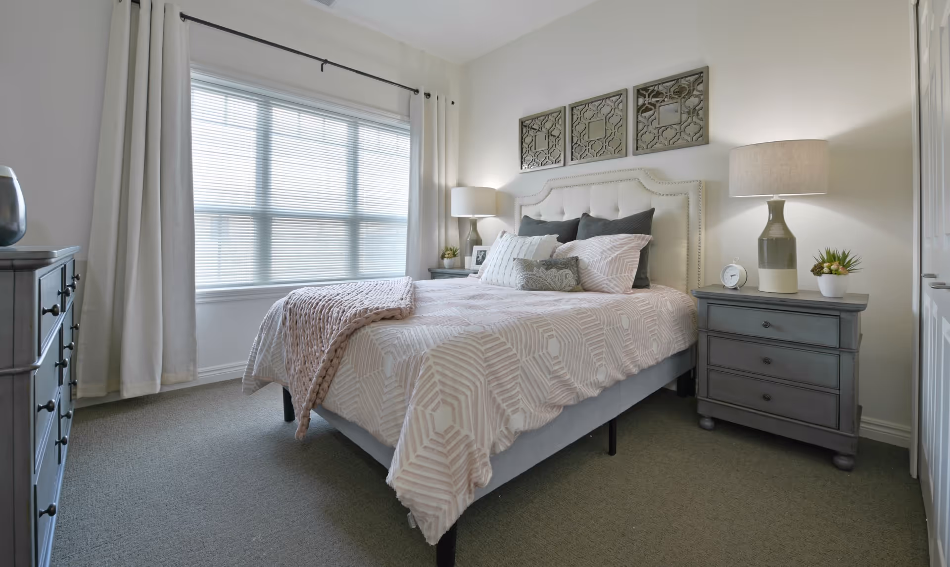 A neatly made bed with patterned bedding and multiple pillows in a bright bedroom. The room has a large window with white curtains, two matching bedside tables with lamps, a small plant, and decorative wall art above the headboard.