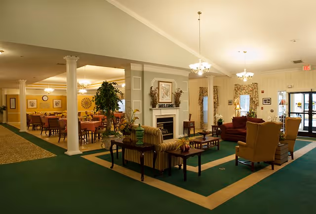 A spacious and warmly lit common area in a senior living facility featuring a seating area with armchairs and sofas around a coffee table, a fireplace with a 'WELCOME' sign above it, and a dining area with multiple tables and chairs in the background. The room has green carpet with beige borders, white columns, and chandeliers hanging from the ceiling.