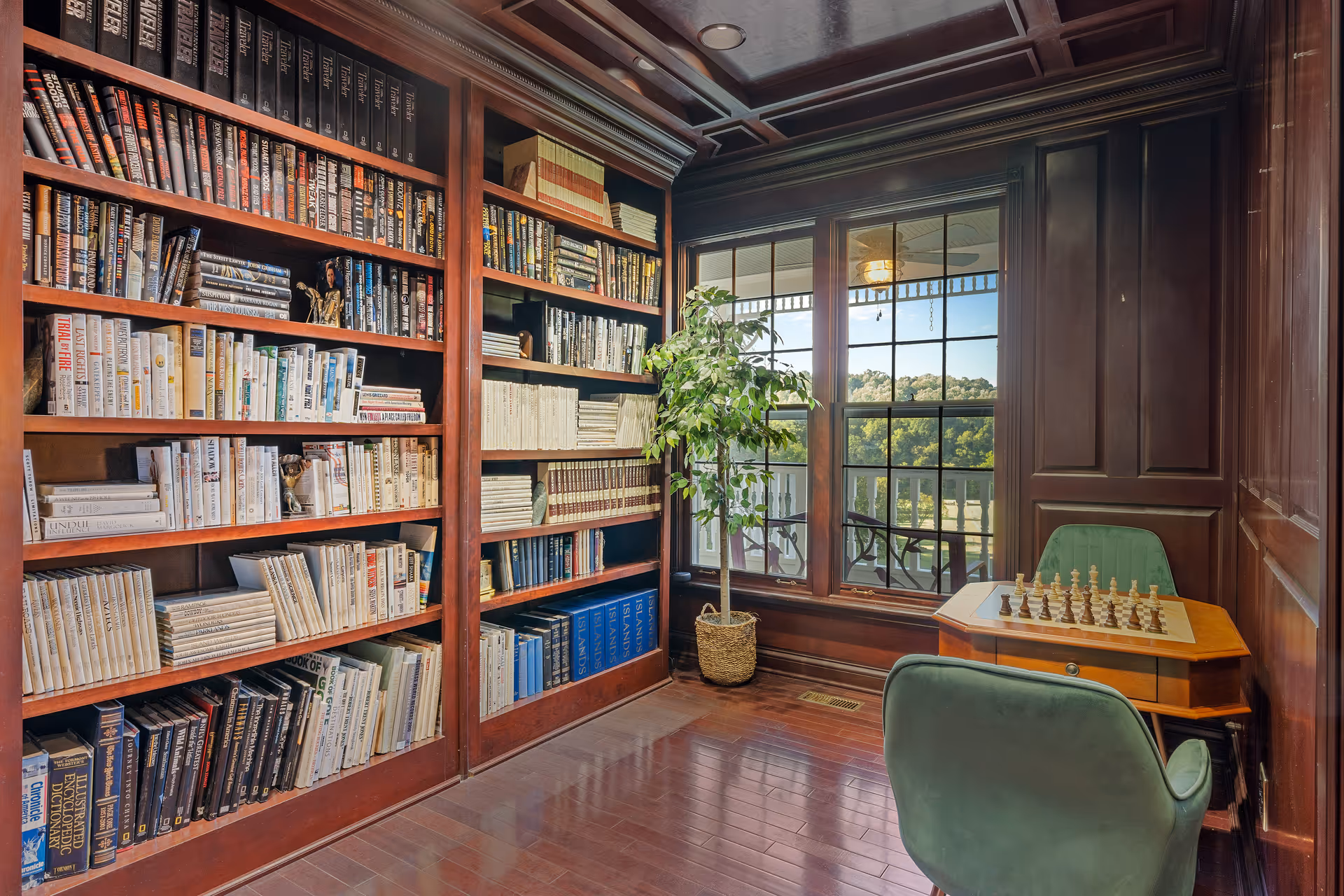 A cozy reading nook with dark wooden bookshelves filled with books, a large window showing a scenic outdoor view, a potted plant, and a small wooden table with a chessboard set up, accompanied by two green upholstered chairs.