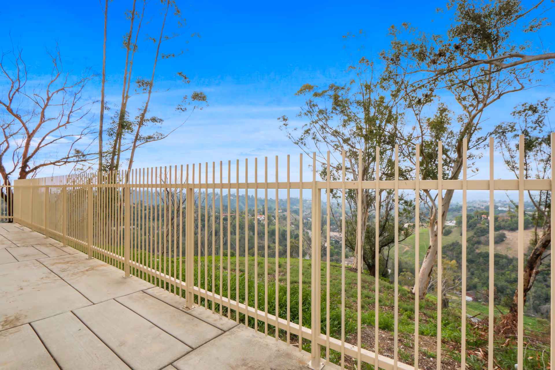 Metal fence along a paved patio overlooking a tree-covered hillside and distant valley.
