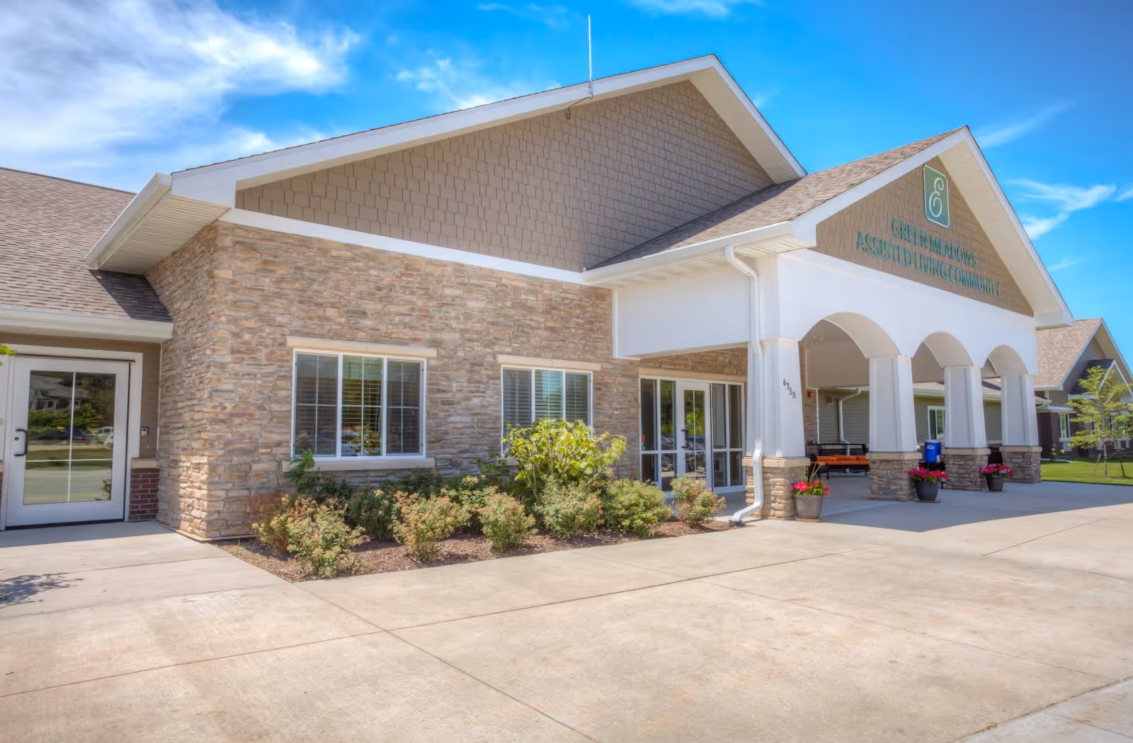 Exterior view of a senior living facility building with stone and siding walls, large windows, a covered entrance with white columns, and a clear blue sky above.
