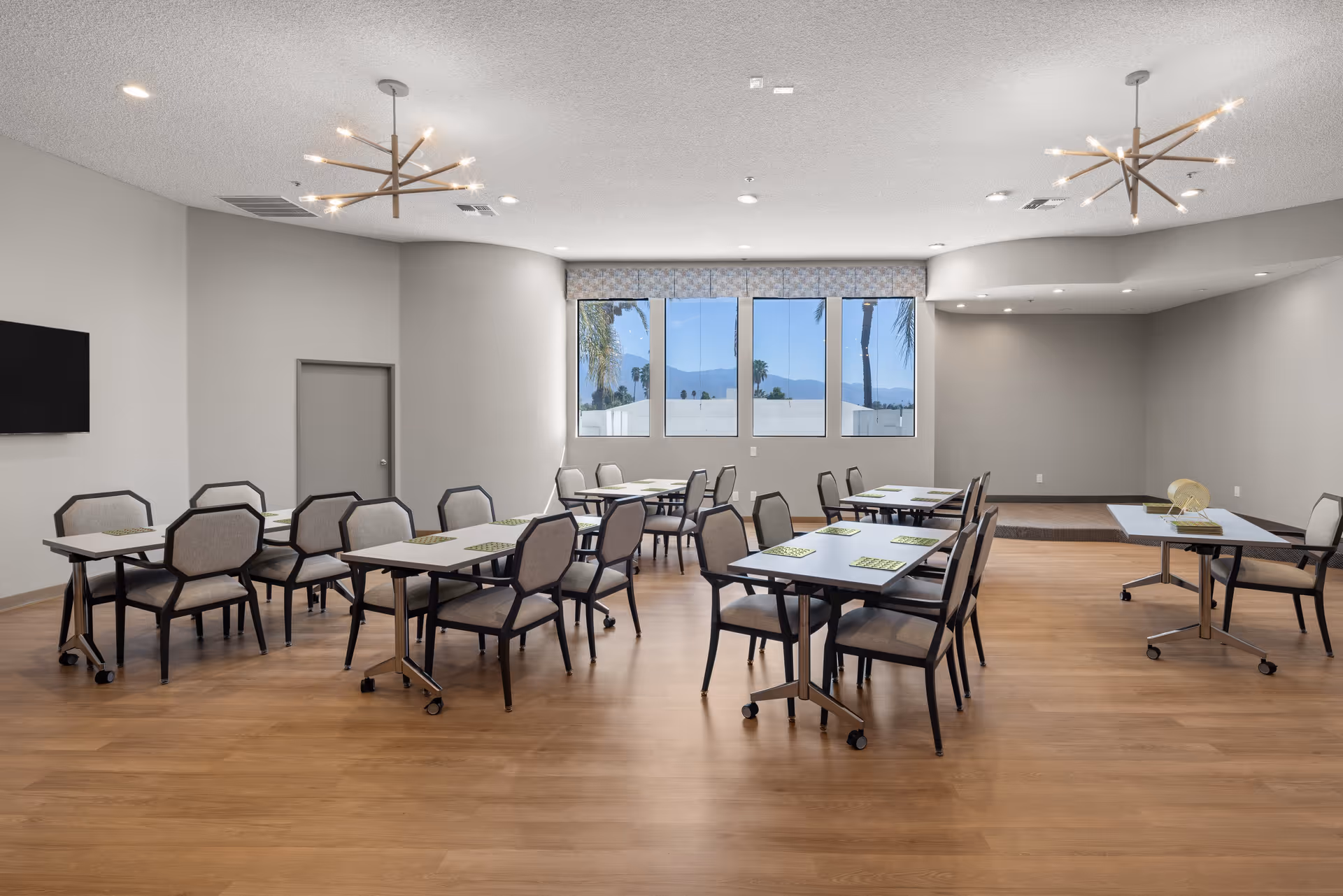 A spacious dining room with multiple tables and chairs arranged neatly on a wooden floor. The room features large windows with a view of palm trees and mountains outside. Modern light fixtures hang from the ceiling, and a flat-screen TV is mounted on one wall.
